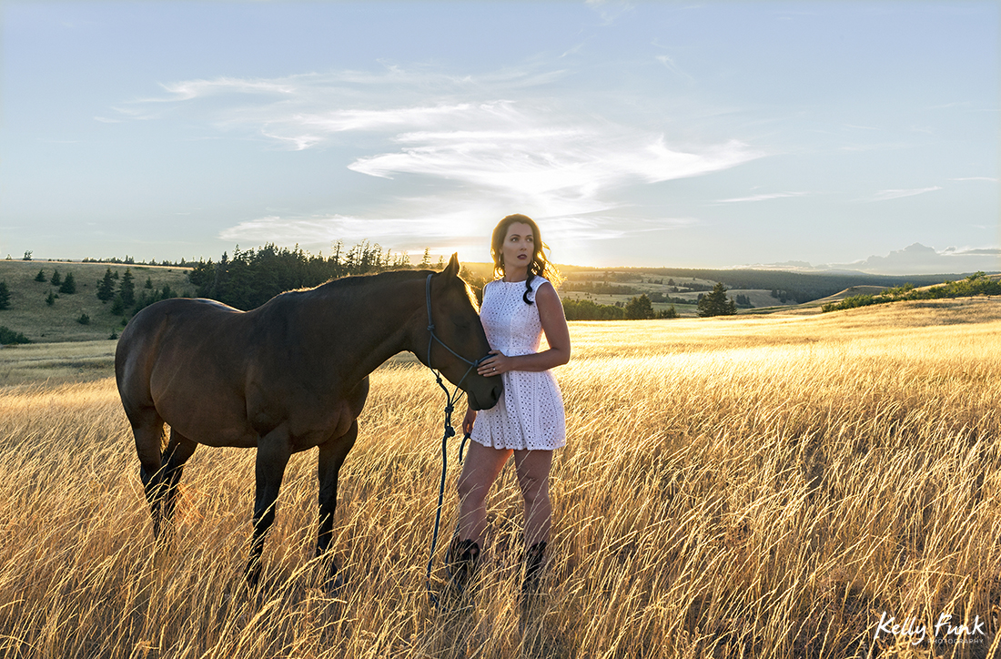 A beautiful woman and her horse pose in a field at sunset for a portrait during sunset on the ranch near Kamloops, British Columbia, Canada